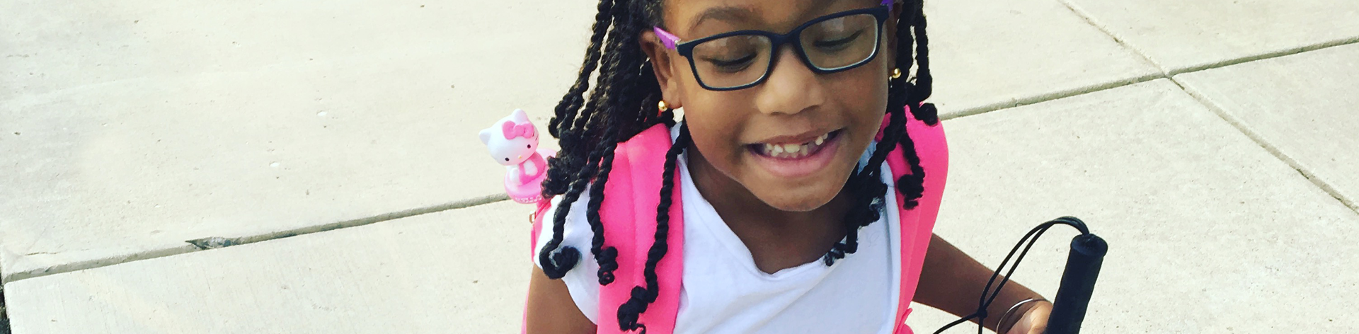 A young blind girl smiles widely. She wears and backpack and her long white cane.